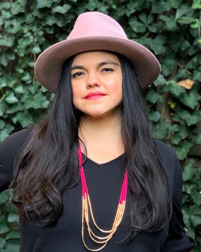 Fair-skinned latin woman with long dark hair wearing a pink wide brimmed hat and a black top smiles at the camera. She is standing in front of green foliage.